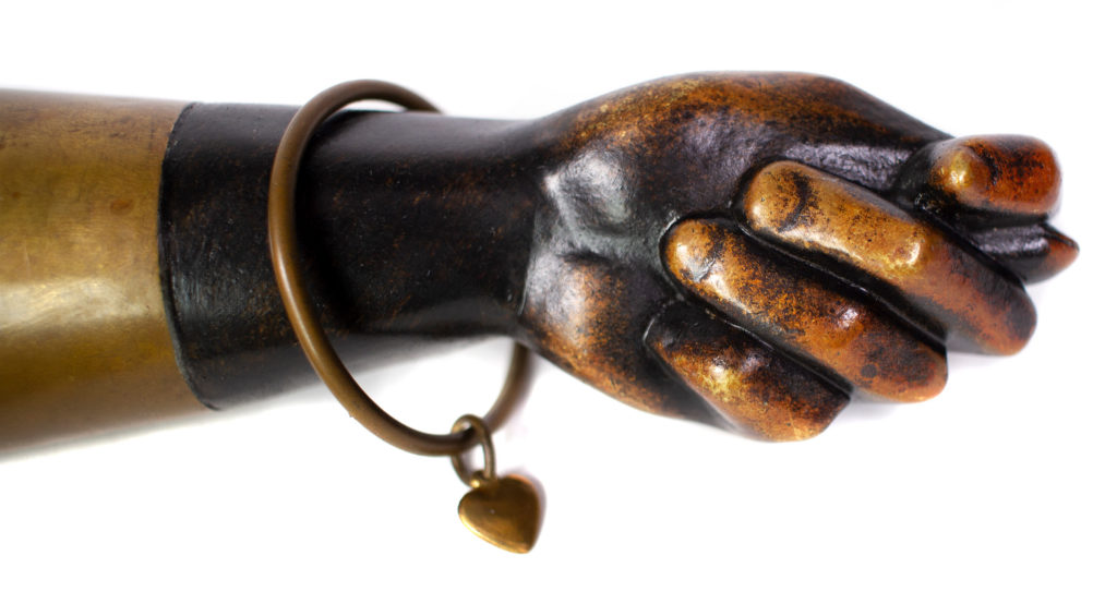 Hand shaped bronze sculpture making a figa gesture with a heart charm bracelet and knot tassel that opens up to reveal a lighter on the top. Marked with "Saks Fifth Avenue" and "Made in Austria."