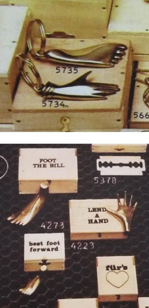 2 Historical catalog photos showing Auböck's use of hand and foot motifs. First photo shows a brass foot keychain under #5735 and brass hand keychain under #5734 sitting on a wooden box. Second photo shows 2 brass foot paperweights and their corresponding wooden boxes with the titles "Foot the Bill" and "best foot forward" and #4273. Also shown is a brass hand paperweight with corresponding wooden box with the title "Lend a Hand" and #4223"