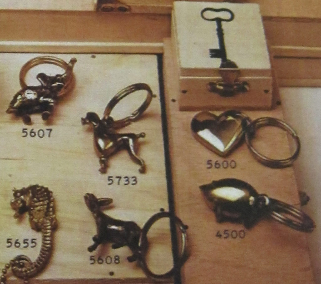 Historical catalog photo showing Auböck's various keychain designs with an elephant under #5607, seahorse under #5655, poodle under #5733, donkey under #5608, heart under #5600, and pig under #4500. All are sitting on top of wooden boxes. 