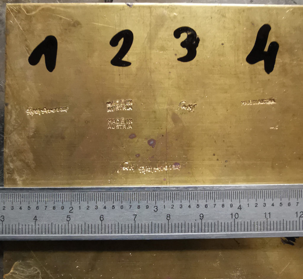Brass sheet with impressed marks from the Auböck estate reading #1 "Fifth Avenue," #2 "Made in Austria" stacked, #3 "Saks," and #4 "Made in Austria" inline with ruler below showing scale.